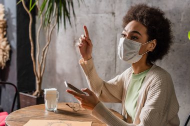 Tıbbi maskeli Afro-Amerikan kadın elinde akıllı telefon ve kafeyi işaret ediyor. 
