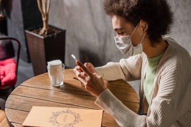 Kafenin yakınlarında akıllı telefon kullanan tıbbi maskeli Afrikalı Amerikalı kadın. 