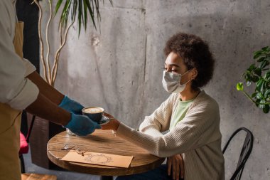 Lateks eldivenli Afro-Amerikan garson kafede maske takan bir kadının yanında kahve tutuyor. 
