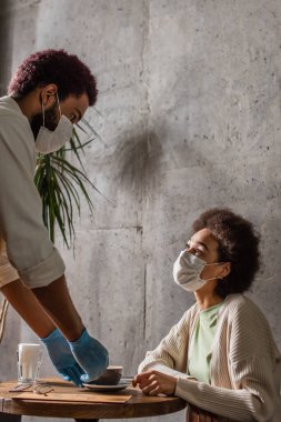 Lateks eldivenli Afrika kökenli Amerikalı garson, tıbbi maskeli müşterinin yanında kapuçino tutuyor. 