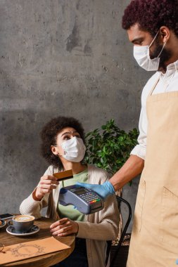 Tıbbi maskede kredi kartı tutan Afrikalı Amerikalı müşteri ödeme terminalini tutuyor. 