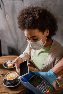 Tıbbi maskeli Afrikalı Amerikalı müşteri kafeteryadaki garsonun yanında akıllı telefonla ödeme yapıyor. 