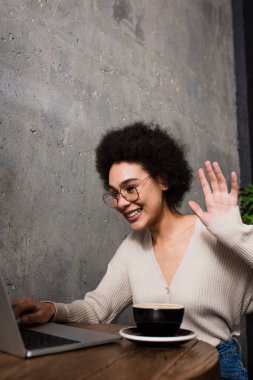 Neşeli Afro-Amerikalı kadın kafedeki kahvenin yanındaki bulanık laptopta video görüşmesi yapıyor. 