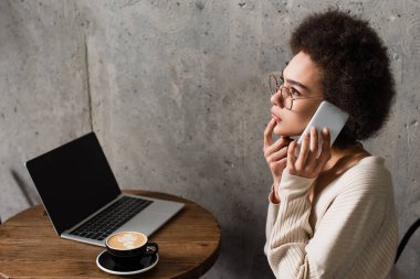 Kafede kahve ve dizüstü bilgisayarın yanında akıllı telefondan konuşan dalgın Afrikalı Amerikalı kadının yan görüntüsü 