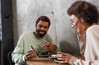 Gülümseyen Afro-Amerikalı adam, bulanık kız arkadaşının yanında akıllı telefonu işaret ediyor ve kafede kahve içiyor. 