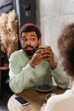 Amazed african american man with coffee looing at blurred girlfriend near smartphone in cafe  clipart
