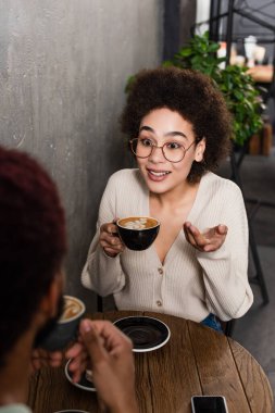 Kahveli, gülümseyen Afro-Amerikan kadın kafede bulanık erkek arkadaşıyla konuşuyor. 
