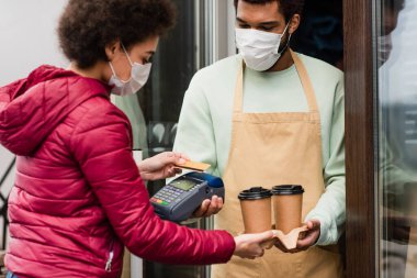 Tıbbi maskeli Afrikalı Amerikalı barista kahve ve ödeme terminalini müşterisinin yanında tutuyor. 