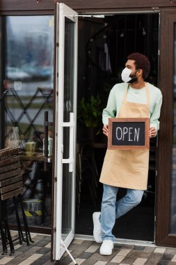 Tıbbi maskeli Afrikalı Amerikalı barista kafeteryada açık harflerle yazı tahtası tutuyor. 
