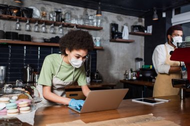African american barista in medical mask using laptop near blurred colleague and coffee machine in cafe  clipart
