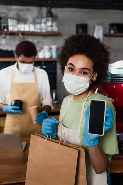 African american barista in medical mask and latex gloves holding cellphone and paper bag in cafe