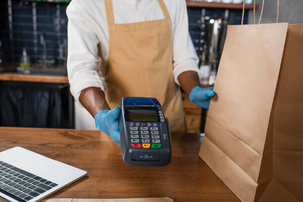 Cropped view of african american barista in latex gloves holding payment terminal and paper bag in cafe 