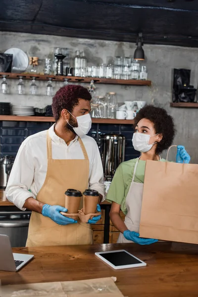 African american baristas in medical masks holding coffee to go and paper bag near gadgets