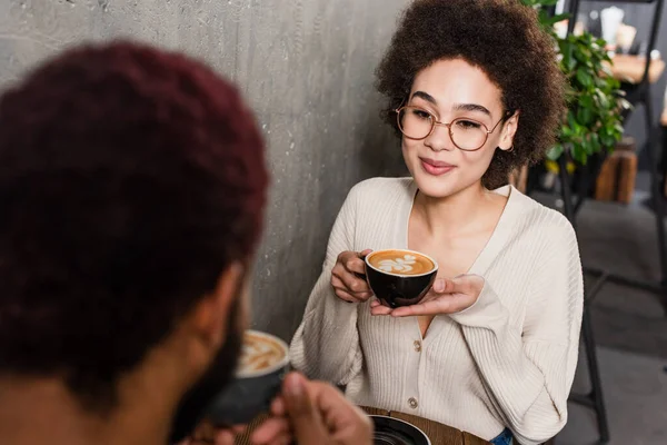 Young african american woman with coffee near blurred boyfriend in cafe