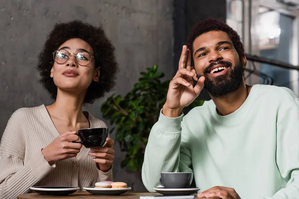 African american man pointing with fingers near girlfriend with coffee and macaroons in cafe