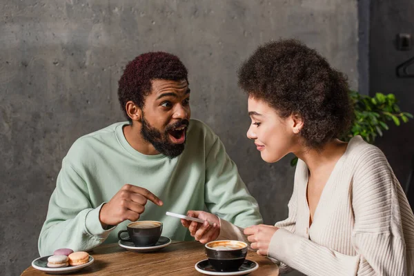 Excited african american man pointing with finger near girlfriend with smartphone and coffee in cafe