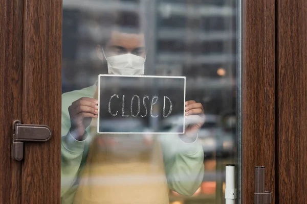 医療用マスクのアフリカ系アメリカ人バリスタで カフェの入り口近くに閉じたレタリングが立っている黒板 — ストック写真