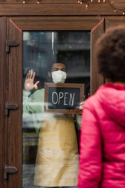 アフリカ系アメリカ人バリスタで医療用マスク手を振って 近くのカフェでオープンレタリングで黒板を保持 — ストック写真