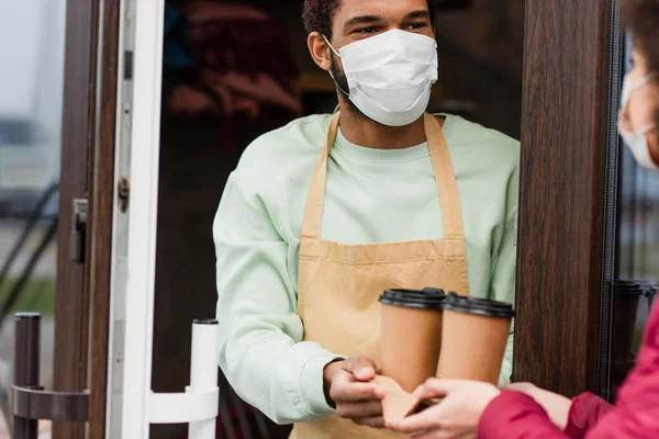 African american barista in medical mask giving coffee to go to blurred client near door of cafe
