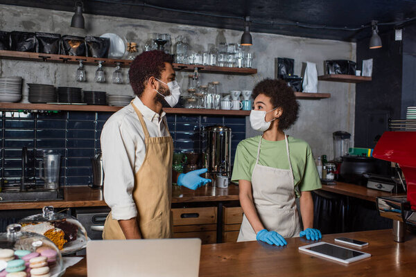 African american baristas in medical masks and latex gloves talking near laptop in cafe 