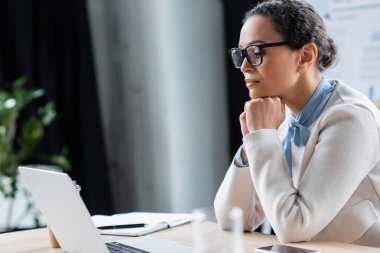 Gözlüklü Afrikalı Amerikalı iş kadını. Bulanık akıllı telefonun yanındaki laptopa bakıyor. 
