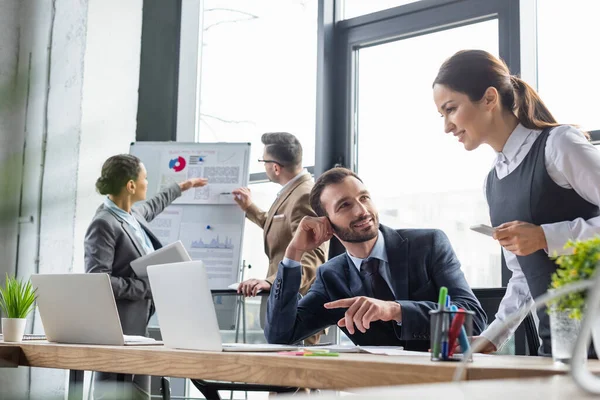 Glimlachende Zakenvrouw Met Smartphone Buurt Van Collega Wijzen Naar Laptop — Stockfoto