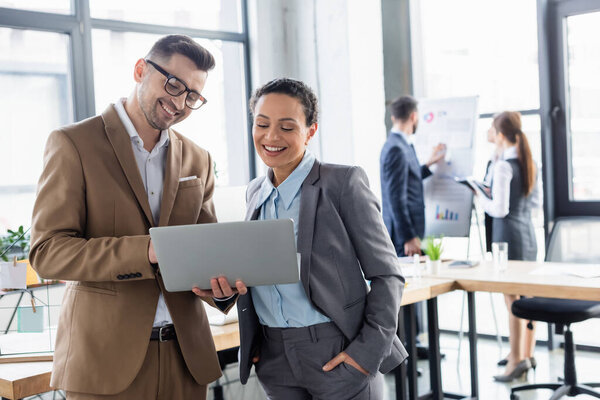 Cheerful businessman holding laptop near african american colleague in office 