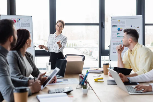 Lächelnde Geschäftsfrau Zeigt Auf Verschwommene Kollegen Und Flipcharts Büro — Stockfoto