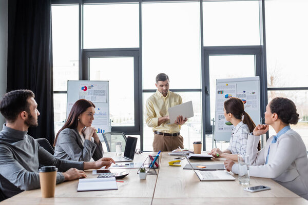 Businessman holding laptop near interracial colleagues and table in office 