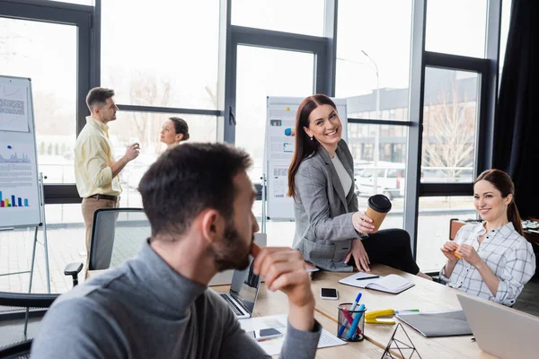 Fröhliche Geschäftsfrau Hält Bei Besprechung Pappbecher Neben Verschwommenem Kollegen — Stockfoto