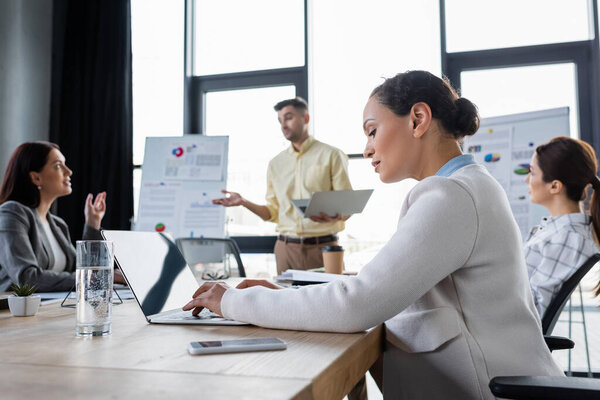 African american businesswoman using laptop near glass of water and blurred colleagues 