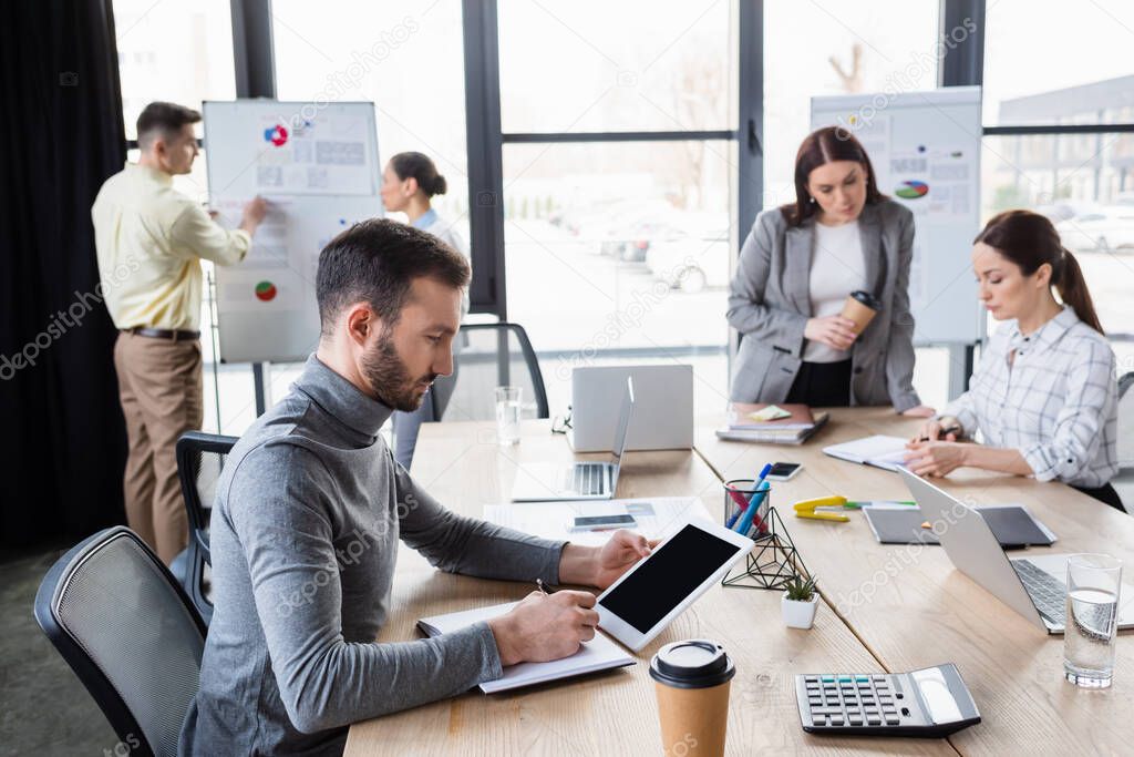 Businessman using digital tablet and writing on notebook near blurred colleagues 