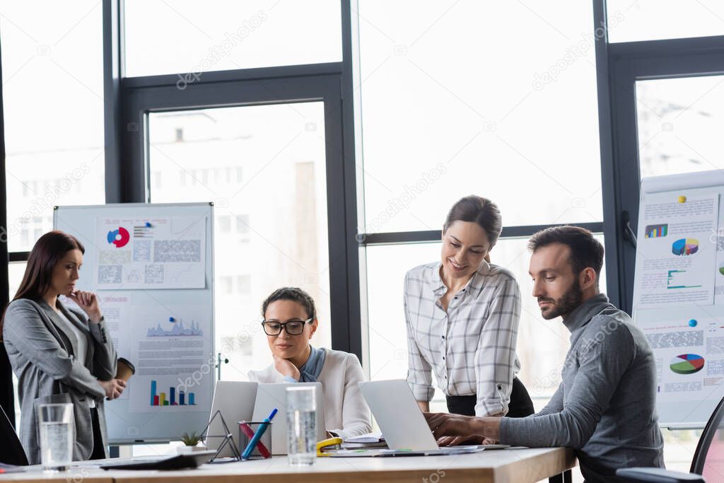 Multiethnic business people using laptops near water and flipcharts in office 