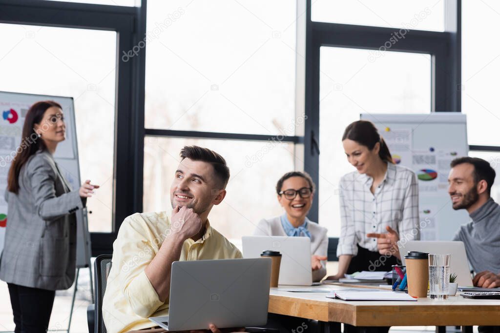 Smiling businessman holding laptop near blurred multiethnic colleagues 