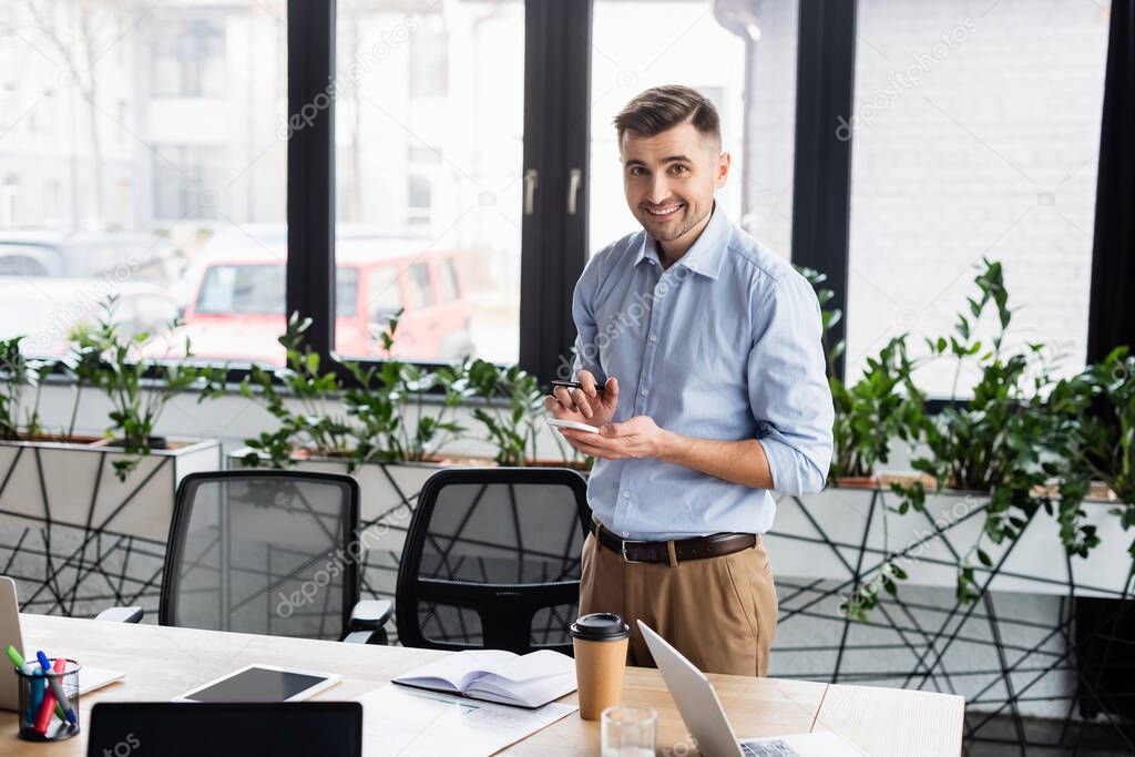 Businessman with pen and smartphone looking at camera near working table 