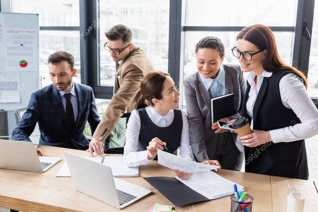 Smiling interracial businesswomen looking at document near businessmen using laptop 