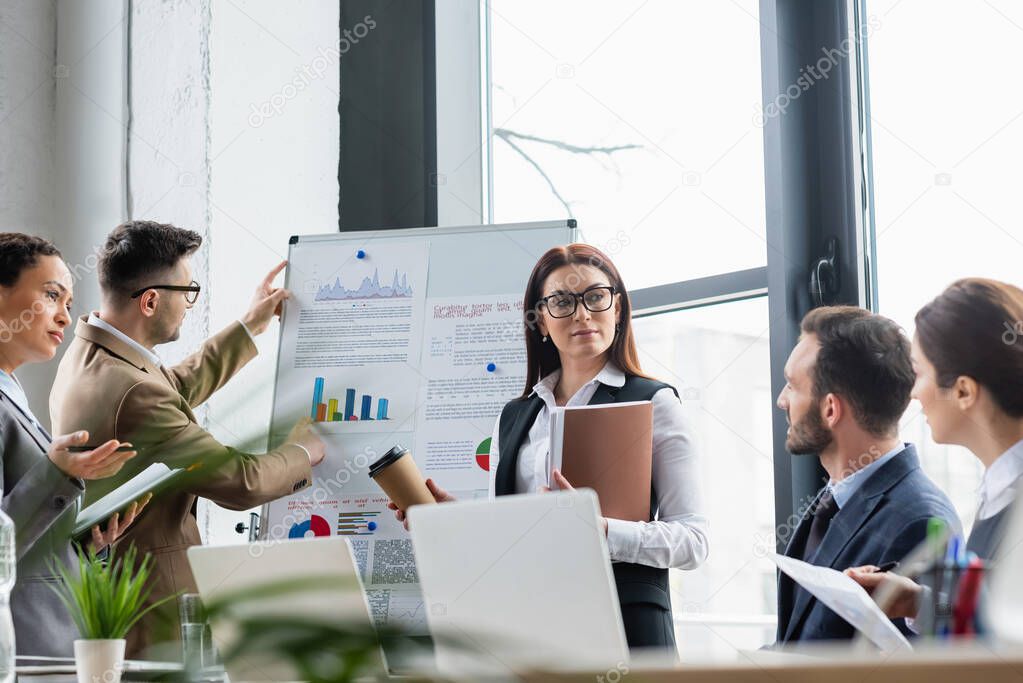 Businesswoman with coffee and paper folder standing near flipchart and multiethnic colleagues 