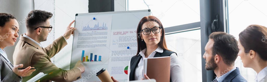 Businesswoman with coffee and papers standing near multiethnic colleagues and flipchart, banner 