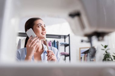 Gülümseyen terzi akıllı telefondan konuşuyor. Ön plandaki dikiş makinesinin yanında. 