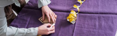 Cropped view of seamstress marking cloth near tape measure, banner  clipart