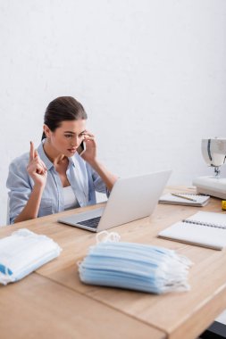 Bilgisayarın, dikiş makinesinin ve tıbbi maskelerin yanında akıllı telefondan konuşan terzi. 