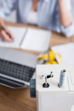 Bulanık terzi ve dizüstü bilgisayarın yakınındaki dikiş makinesinin kırpılmış görüntüsü 