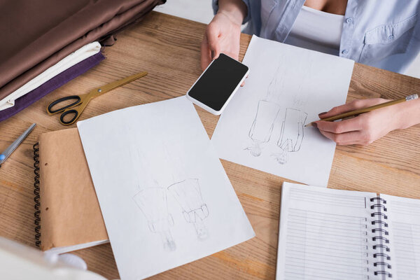 Cropped view of seamstress with smartphone drawing near cloth on table 