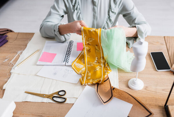 Cropped view of fabric in hands of seamstress working near smartphone and mannequin 
