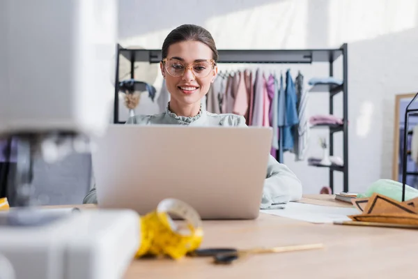 Lächelnde Näherin Mit Laptop Der Nähe Verschwommener Nähmaschinen Und Muster — Stockfoto