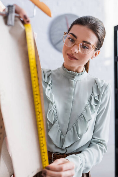 Designer in eyeglasses measuring sewing pattern on blurred foreground 