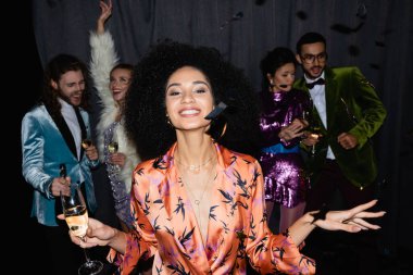 Gülümseyen Afrikalı Amerikalı kadın gri arka planda ırklar arası arkadaşlarının yanında bir kadeh şampanyayla dans ediyor.