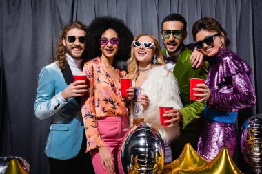 Güneş gözlüklü, gri arka planda plastik bardaklarla duran ırklar arası gülümseyen arkadaşlar.