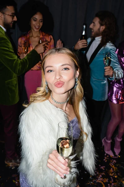 young adult woman pouting lips with glass of champagne on party with interracial friends on grey background