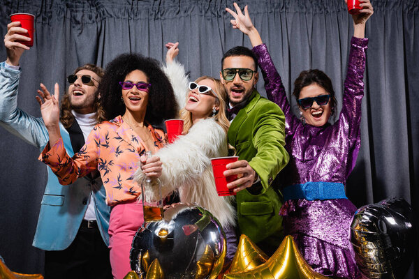 smiling interracial friends in sunglasses having party on grey background